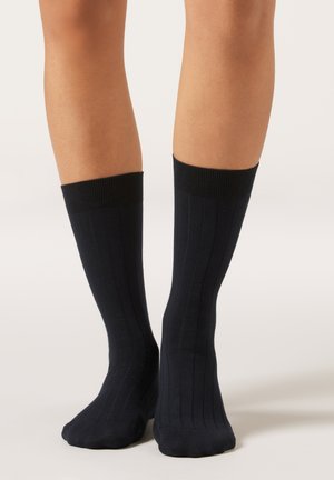 Benedenbenen met zwarte geribbelde sokken tot halverwege de kuit die een effen lichte achtergrond raken, met een dikke boord bovenaan.