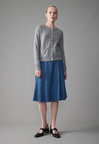Grå stickad kofta med knappar framtill och fickor, matchad med en knälång blå denimkjol. Modell bär svarta blockklackskor.