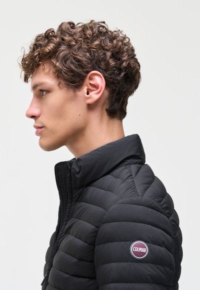 Jeune homme aux cheveux bouclés portant une veste matelassée noire à col haut avec un logo rond Colmar sur la manche, tourné vers la gauche.