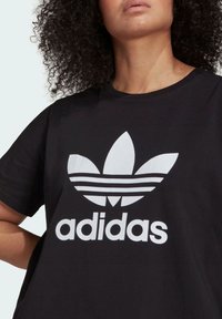 Černé bavlněné tričko s velkým bílým logem Adidas trefoil a textem pod ním; kulatý výstřih a krátké rukávy.