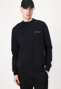 Fekete pamut sweatshirt, klasszikus környakú kivitelben, bordázott mandzsettákkal és két kis fehér márkájú logóval a mellrészen.