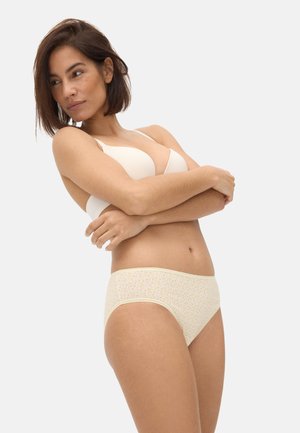 Mujer con cabello corto y castaño, vestida con un brasier beige y ropa interior de color amarillo claro con estampado floral, de pie con los brazos cruzados.