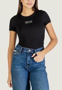 T-shirt nera in cotone con maniche corte e scollo rotondo, con un logo bianco "TOMMY JEANS" ripetuto sul petto.