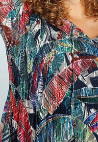 Nainen, jolla on kihara hius, pukeutuneena moniväriseen mekkoon, jossa on voimakkaat, abstraktit lehtikuviot punaisessa, sinisessä, vihreässä ja valkoisessa sävyssä.