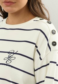Vit randig topp med marinblå linjer, med rund halsringning och dekorativa knappar på axeln. Tyg med textur och logodetalj.