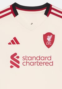 Kreminė džersio maikutė su raudonais akcentais, turinti V formos iškirpimą, tris raudonas juostas ant pečių, Liverpulio FC emblema ir "Standard Chartered" logotipas.