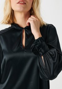 Blusa in satin nero con scollatura a chiave, maniche a sbuffo e dettagli con bottoni nei polsini. Texture liscia, design elegante senza motivi.