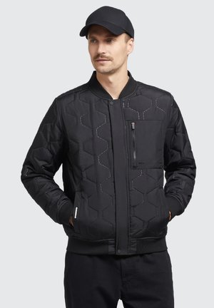 Schwarze gesteppte Bomberjacke mit hexagonalem Muster, Reißverschlusstasche, gerippten Manschetten und Kragen. Getragen mit einer passenden schwarzen Mütze.