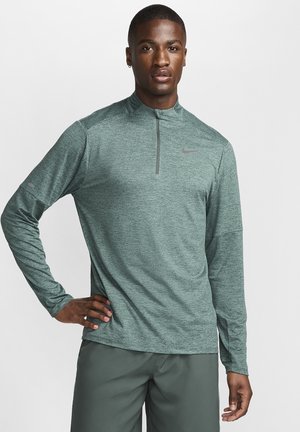 Vihreä pitkähihainen puolivetoketjullinen paita teksturoidusta kankaasta, jossa on hienovarainen heather-kuvio. Paidan rintamalla on harmaa Nike-logo ja käsivarren kohdalla yksityiskohta.
