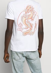 Bílý bavlněný tričko s podrobným grafickým vyobrazením oranžového draka na zadní straně, doplněné lotosovým květem a stylizovaným textem.