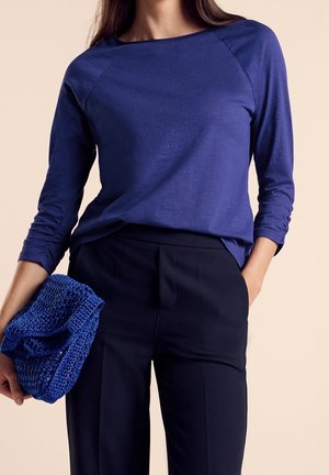 Kobieta ubrana w długi niebieski top z długim rękawem i czarne spodnie, trzymająca w jednej ręce złożoną niebieską dzianinową kopertówkę.