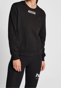Sweat-shirt noir à manches longues, avec poignets et ourlet côtelés, arborant un logo blanc "HUMMEL" sur la poitrine. Tissu doux avec une coupe classique.