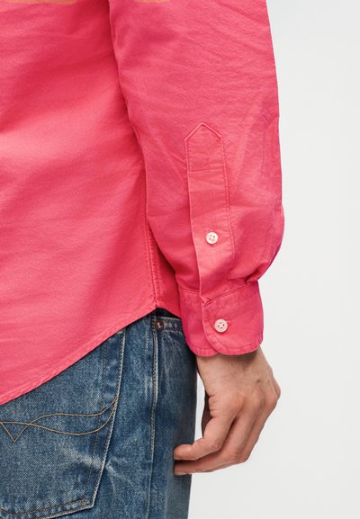 Άνδρας που φοράει ροζ πουκάμισο με μακριά μανίκια και κουμπωτό μανσέτα, μπλε τζιν τζιν, με το χέρι ακουμπισμένο στο πλάι, μπροστά σε απλό λευκό φόντο.
