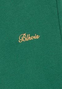 Un tissu vert présente une texture lisse avec un petit logo brodé en or "Blkvis". Le design est simple et minimaliste.