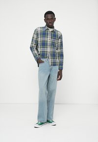 Chemise à carreaux verte, bleue et blanche, boutonnée, portée avec un jean droit bleu clair. Des baskets vertes et blanches complètent le look.
