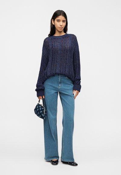 Pull en maille bleu marine avec manches larges, coupe décontractée ; assorti à un jean évasé bleu clair et un petit sac à motif. Des ballerines noires complètent la tenue.