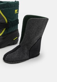 Forro de bota de fieltro gris con un acento de piel negra, con forma curva y textura suave. Se puede ver la marca Sorel y una bota de nylon adyacente.