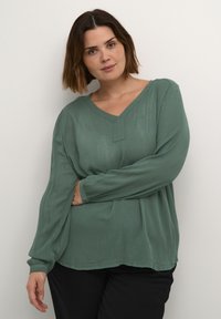 Blouse verte à manches longues en tissu léger avec un décolleté en V et une surface texturée. Présente une coupe décontractée et un léger plissage à l'avant.