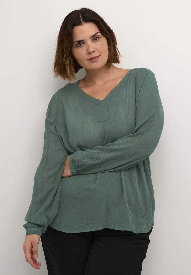 Blouse verte à manches longues en tissu léger avec un décolleté en V et une surface texturée. Présente une coupe décontractée et un léger plissage à l'avant.