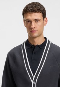 Tummanharmaa neuletakki valkoisilla reunuksilla, nappikiinnitys ja alla poolopaita. Ominaisuuksina teksturoitu kangas ja vasemmalla puolella logo.