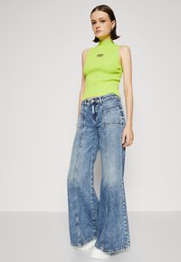 Neon green ribbed sleeveless top with a round logo detail, paired with light blue wide-leg denim jeans, and white platform sneakers.