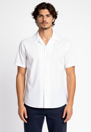 Uomo con capelli castani di media lunghezza che indossa una camicia bianca a maniche corte abbottonata e pantaloni scuri su sfondo bianco.