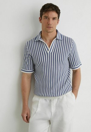 Homme portant une chemise à manches courtes rayée verticalement bleu et blanc et un pantalon blanc, debout devant un mur clair uni.