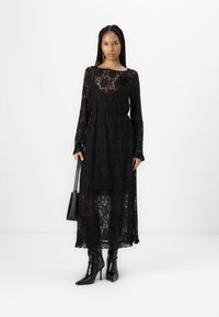 Μαύρο φόρεμα με δαντέλα και μακριά μανίκια, λουλουδάτο σχέδιο, διάφανη επικάλυψη και εφαρμοστός σχεδιασμός στη μέση. Συνδυάζεται με μαύρα μποτάκια με τακούνι και μια μαύρη τσάντα.