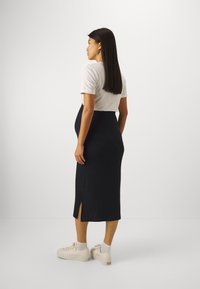 Robe maxi noire avec une fente sur le côté, associée à un haut à manches courtes blanc et des baskets beiges. Modèle de profil, mettant en valeur l'ajustement de la tenue.