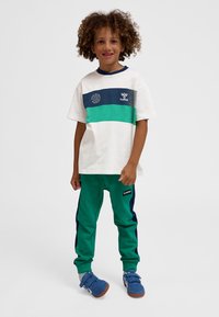 Fiatal, göndör hajú gyermek fehér, kék és zöld sportfelszerelésben, állva és mosolyogva egy sima fehér háttér előtt.