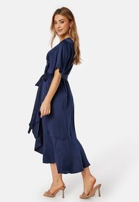 Middellange navyblauwe wikkeljurk van satijn met korte mouwen, een tailleband en een gelaagde ruffle zoom, gecombineerd met naaktkleurige hoge sandalen.