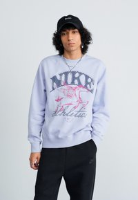 Fiatal személy fekete sapkát, világos lila Nike pulóvert szárnyszöcske mintával és fekete nadrágot visel, az egyik kezét zsebében tartva áll.