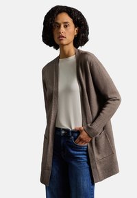 Długi, otwarty kardigan z miękkiego brązowego materiału, z prążkowanymi mankietami i dwiema przednimi kieszeniami. W połączeniu z białą bluzką i niebieskimi dżinsami.