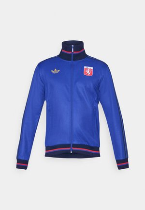 Plava sportska jakna s visokim ovratnikom, zatvaranjem na zip, zlatnim detaljima, bočnim prugama i klubskim logotipom na prsima. Ima rebraste manžete i donji rub.