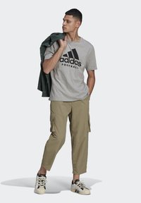 T-shirt in cotone grigio con logo "adidas Football" nero, abbinato a una giacca camicia verde, pantaloni cargo beige e sneakers nere/bianc.