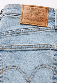 Light blue denim jeans with a brown leather patch featuring embossed details and orange stitching along the seams. Back pocket with curved design.