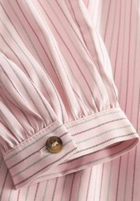 Světle růžová košile s vertikálními pruhy v černé a tyrkysové barvě, se stahovaným manžetem a knoflíkem v kontrastní barvě.