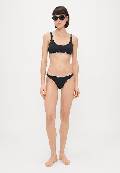 Juoda bikinio komplektas su trumpa palaidine ir žemais apatiniais, akcentuotas kontrastinga balta siūle prie kraštų. Lygus audinio paviršius.