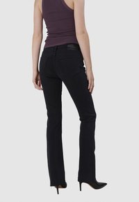 Jean noir évasé avec une texture lisse, doté d'une étiquette en cuir à l'arrière de la taille et de deux poches arrière. Porté avec un débardeur violet.