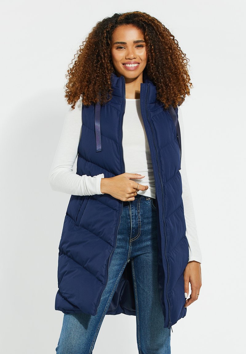 Smilende kvinde med krøllet hår iført en navyblå, polstret ærmeløs vest, hvid skjorte med lange ærmer og blå jeans mod en hvid baggrund.