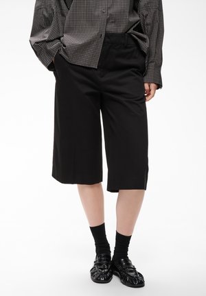 Persoon draagt zwarte knielange shorts, zwarte sokken, glanzende zwarte loafers en een los geruit overhemd met lange mouwen met handen in de zakken.
