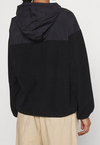 Černá mikina s kapucí s kontrastními matnými a lesklými texturami, která má stahovací kapuci a volnější střih.