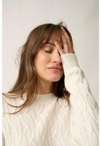 Kremowy sweter z splotem w warkocze, okrągłym dekoltem i długimi rękawami. Modelka dotyka czoła, prezentując delikatne fale we włosach.