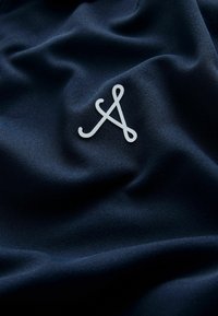 Tissu bleu marine avec une texture lisse, arborant un logo blanc fluide dans le coin inférieur droit. Le matériau semble doux et luxueux.