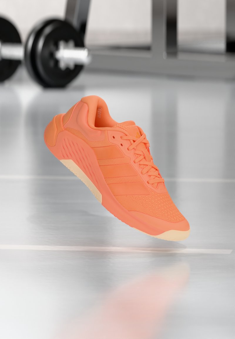 Πορτοκαλί αθλητικό παπούτσι με ανθεκτικό επάνω μέρος από αεριζόμενο δίχτυ, υφασματικές λεπτομέρειες και τρεις ρίγες στο πλάι. Διαθέτει ελαφριά σόλα από καουτσούκ.