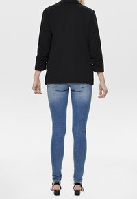 Blazer negro con un diseño estructurado, mangas enrolladas, combinado con jeans ajustados de color azul claro. El conjunto presenta un hardware mínimo y una textura suave.