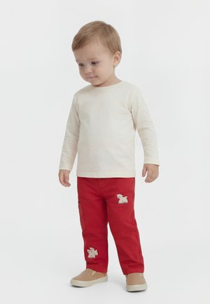 Dijete stoji, odjeveno u kremastu majicu dugih rukava, crvene hlače s aplikacijama pasa i bež cipele na izvlačenje, na jednobijeloj pozadini.