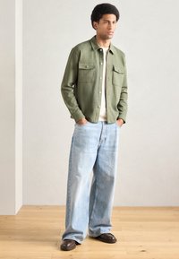 Camicia verde con bottoni e due tasche sul petto, abbinata a jeans azzurri a gamba larga e scarpe marrone scuro su pavimento in legno.