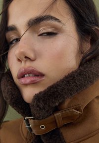 Collier en shearling marron avec une sangle en cuir et une boucle. Gros plan sur un visage avec une peau claire, des lèvres pulpeuses et des cheveux foncés aux légères ondulations.