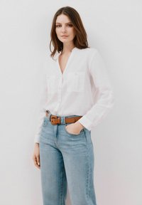 Camicia bianca con colletto a V, due tasche sul petto e maniche lunghe. Abbinata a jeans a vita alta di colore blu chiaro e una cintura marrone.